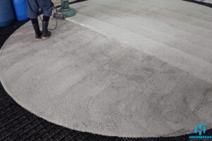 Profissional da Higienizzar operando equipamento de extração profunda em um tapete redondo, mostrando o contraste nítido entre as fibras sujas e a área revitalizada. Serviço de alta tecnologia da nossa lavanderia de tapetes em Itapema.
